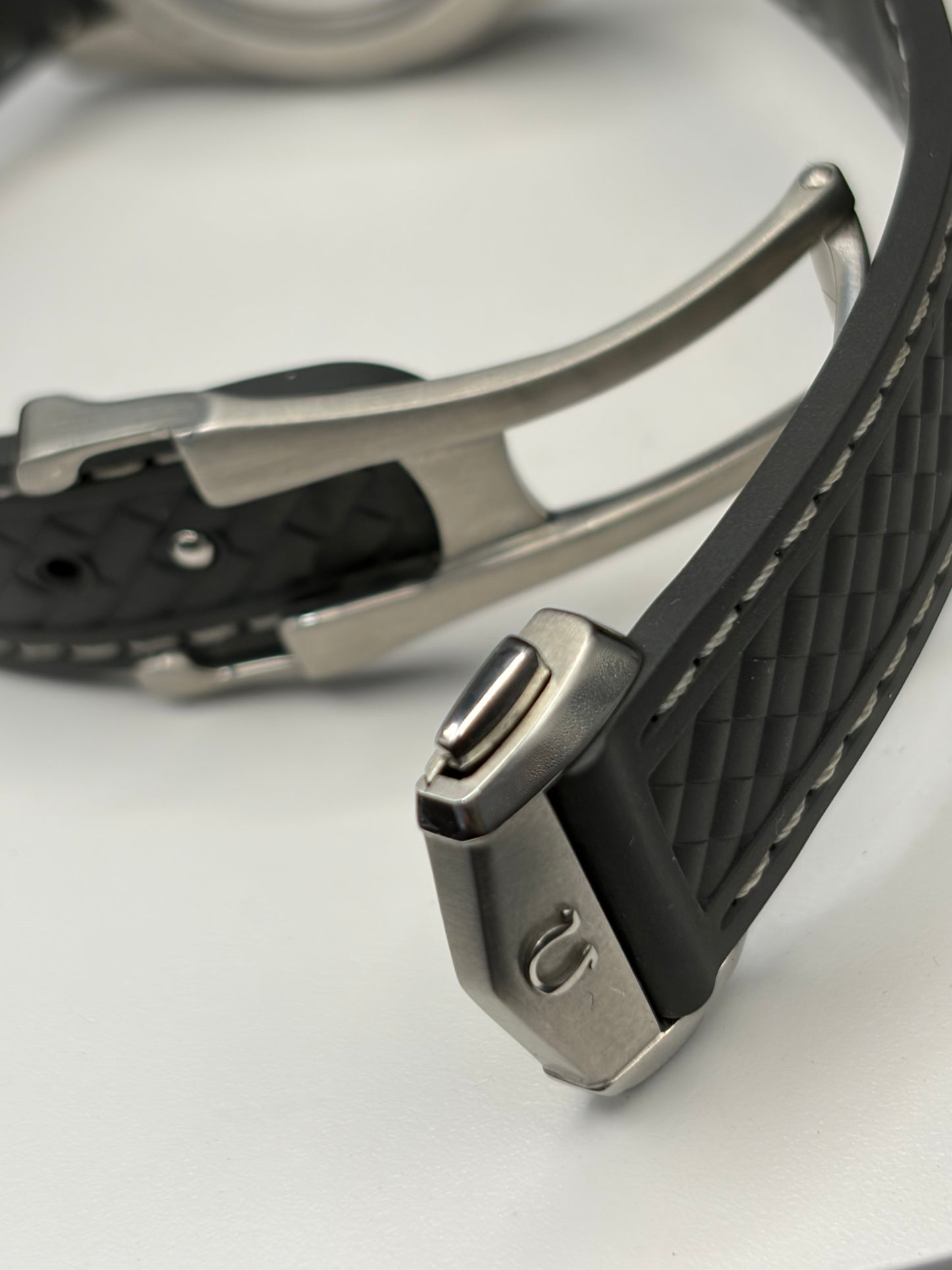 Close-up of a watch strap with a black textured silicone band and a silver-toned metal buckle on a white surface.