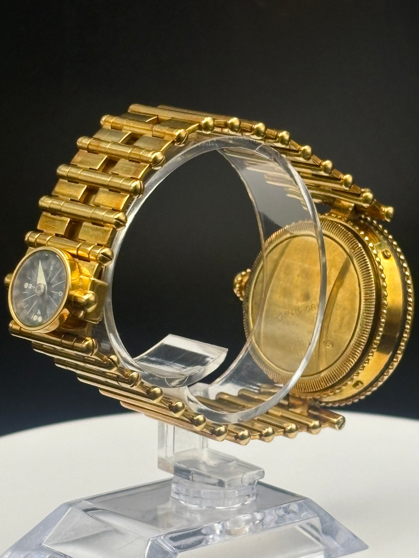 A gold-tone watch with a matching link bracelet, displayed on a clear acrylic stand against a dark background. The watch face is visible on the left, and the back casing is visible on the right.