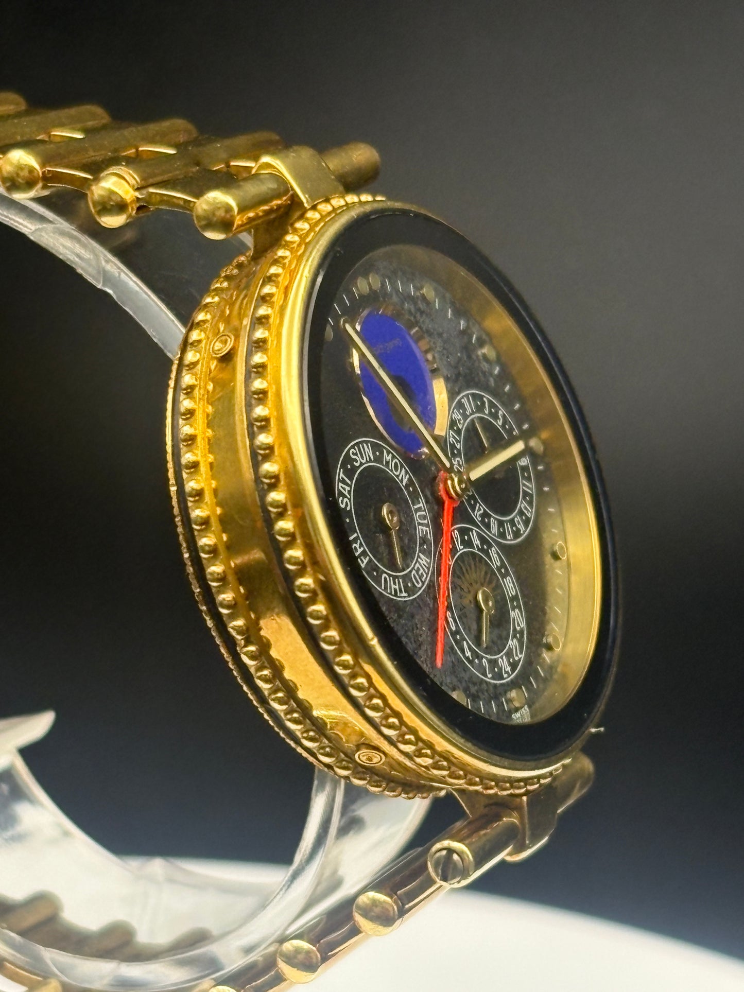 A close-up of a gold-tone wristwatch with a decorative bezel, gold link bracelet, and a black dial featuring moonphase indicators, subdials, and a blue moon phase accent.