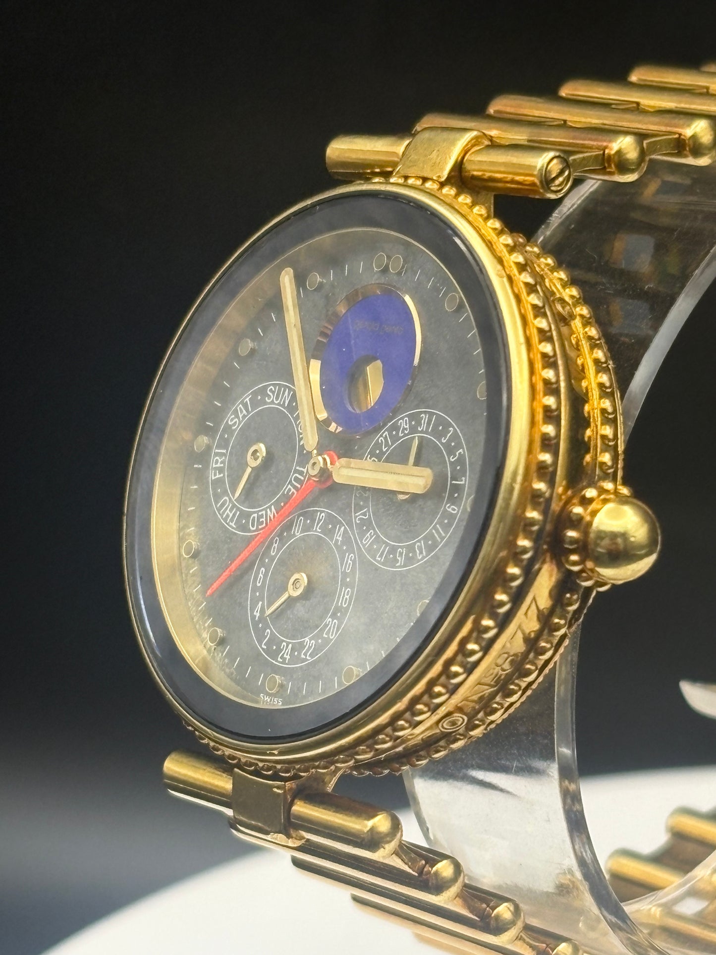 A close-up of a gold-tone wristwatch with a dark gray dial, multiple subdials, a blue moon phase indicator, and a gold link bracelet, displayed on a clear stand against a dark background.