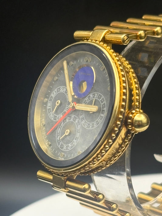A close-up of a gold-tone wristwatch with a dark gray dial, multiple subdials, a blue moon phase indicator, and a gold link bracelet, displayed on a clear stand against a dark background.