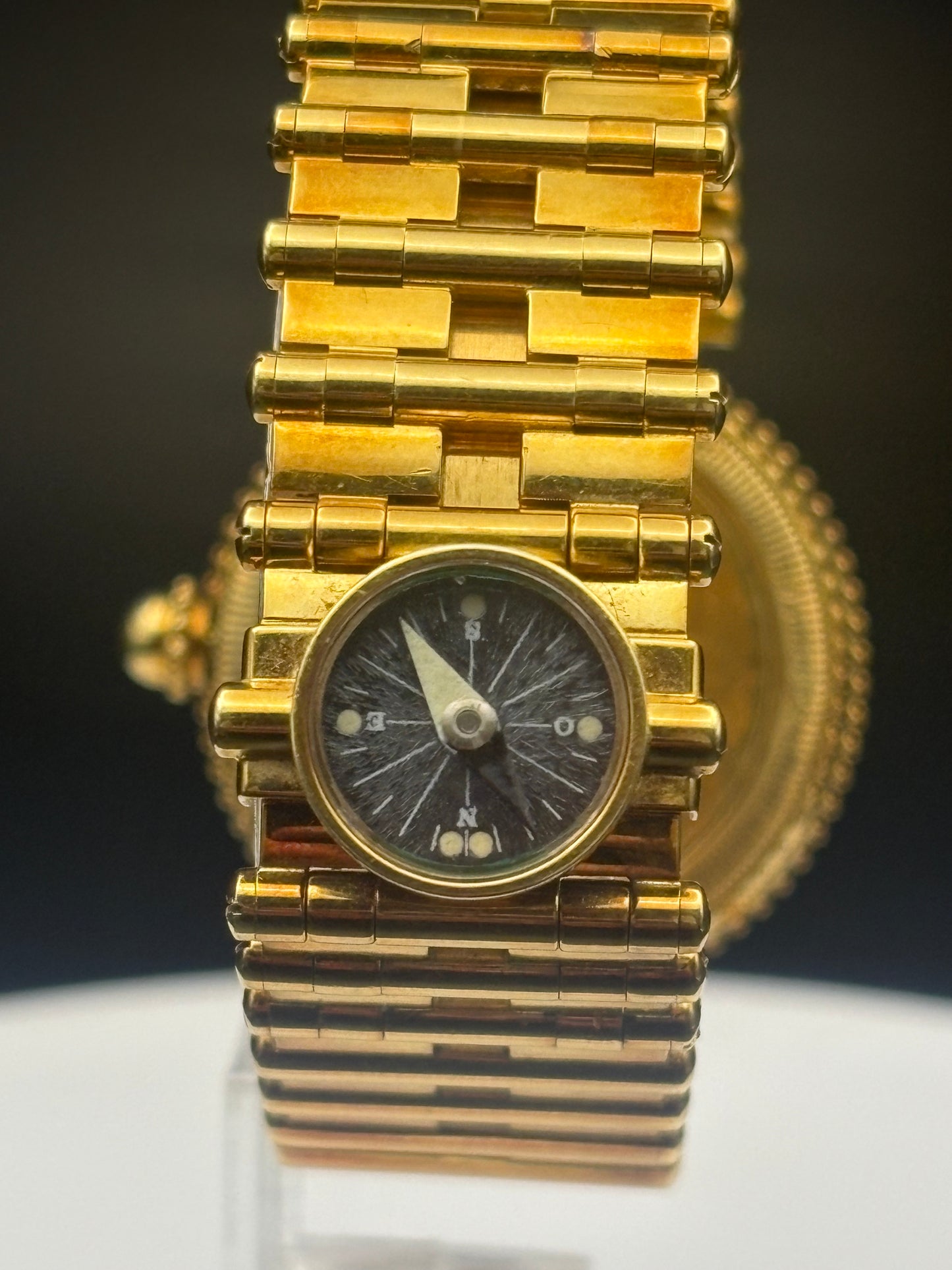 A vintage gold-tone wristwatch with a multi-link bracelet and a dark dial featuring a compass rose, displayed on a reflective white surface against a dark background.