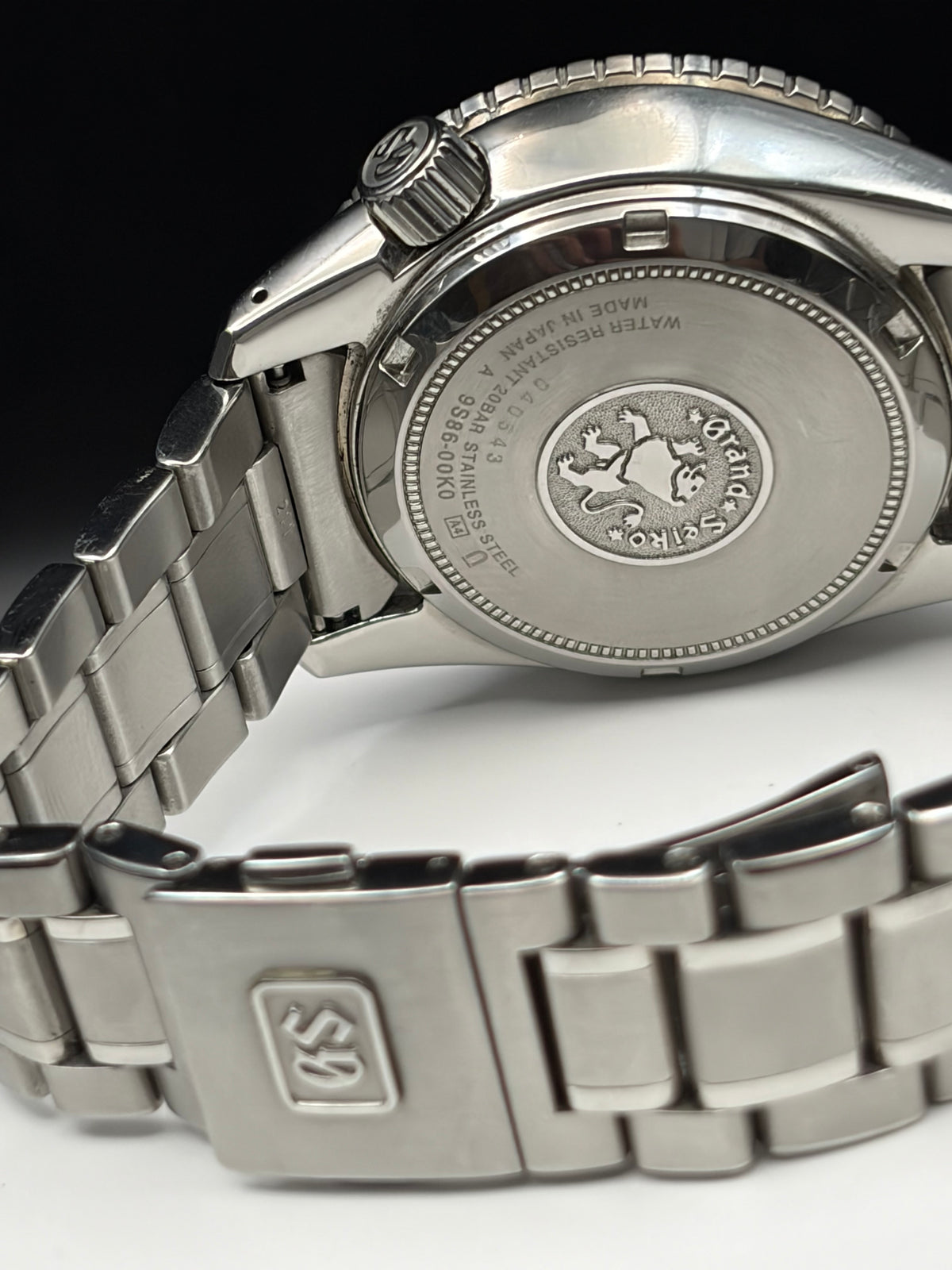 Close-up of the back of a stainless steel wristwatch with a metal link bracelet, showing engraved branding and a clasp with a logo, photographed against a dark and light background.