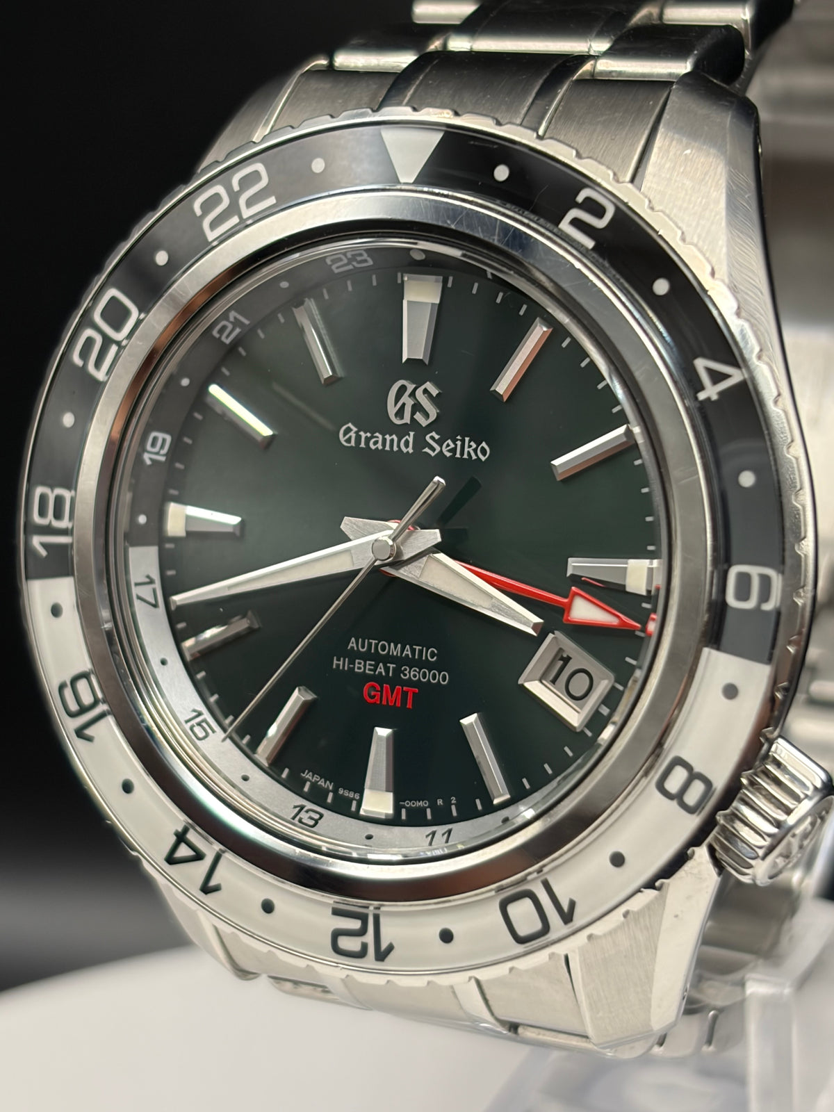 A close-up shot of a Grand Seiko wristwatch with a stainless steel bracelet, featuring a dark green dial, a black bezel with white numerals, and a red GMT indicator hand.