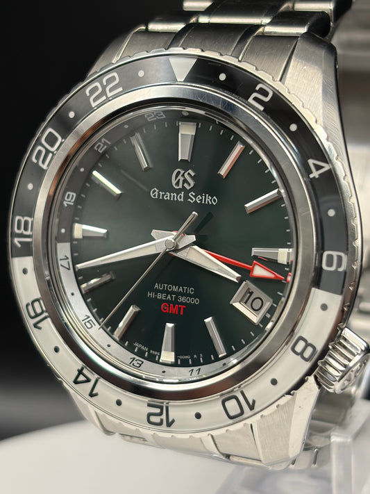 A close-up shot of a Grand Seiko wristwatch with a stainless steel bracelet, featuring a dark green dial, a black bezel with white numerals, and a red GMT indicator hand.