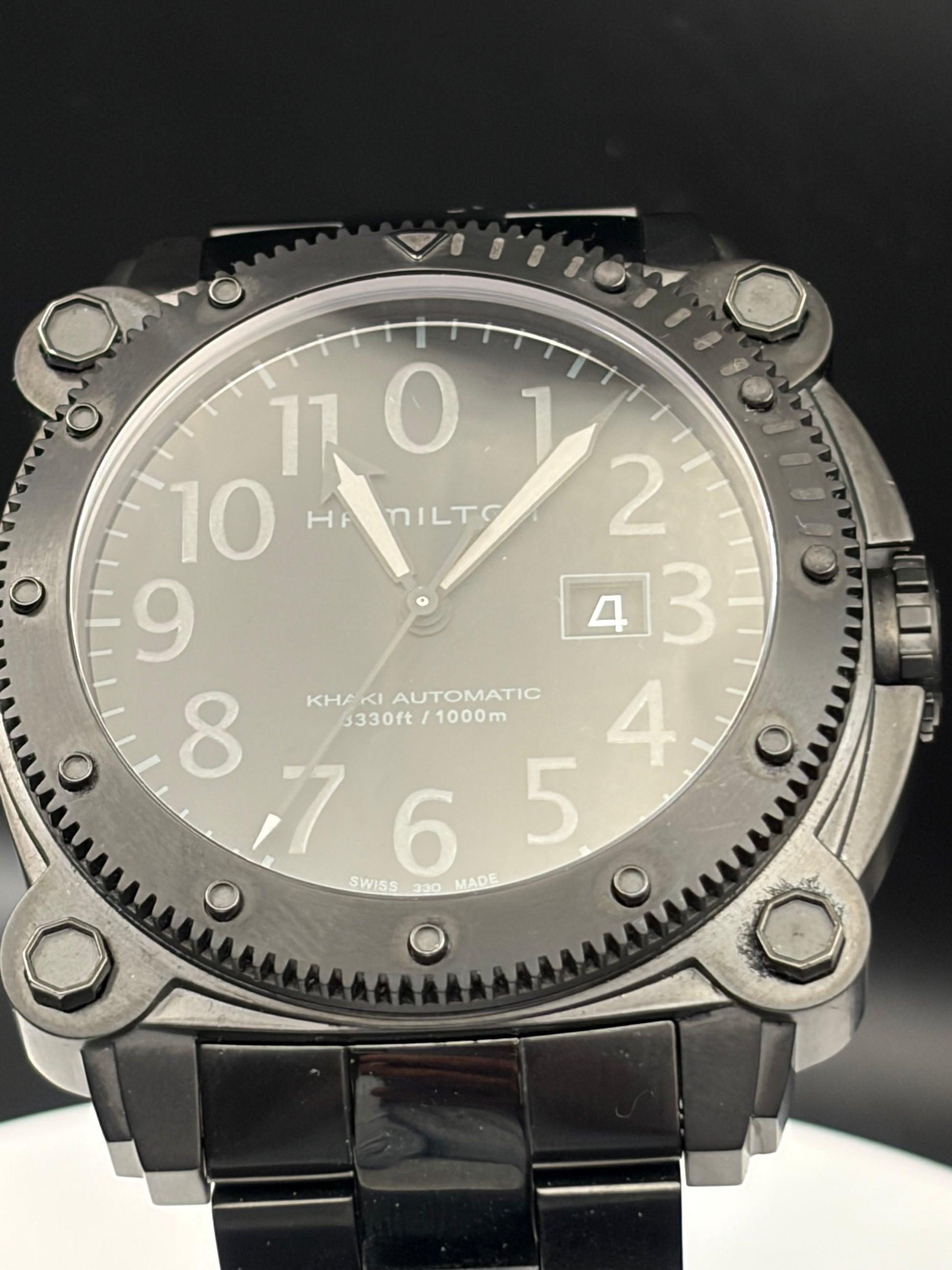 Hamilton Khaki Automatic Observatory watch with a dark gray dial, a textured dark bezel, and a black link bracelet, shown in a close-up shot against a dark background.