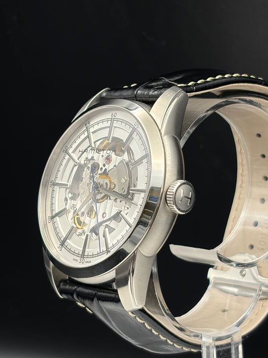 A luxury skeleton watch with a silver stainless steel case, a white skeleton dial showing the inner movement, and a black leather strap with white stitching, displayed on a clear stand against a black background.