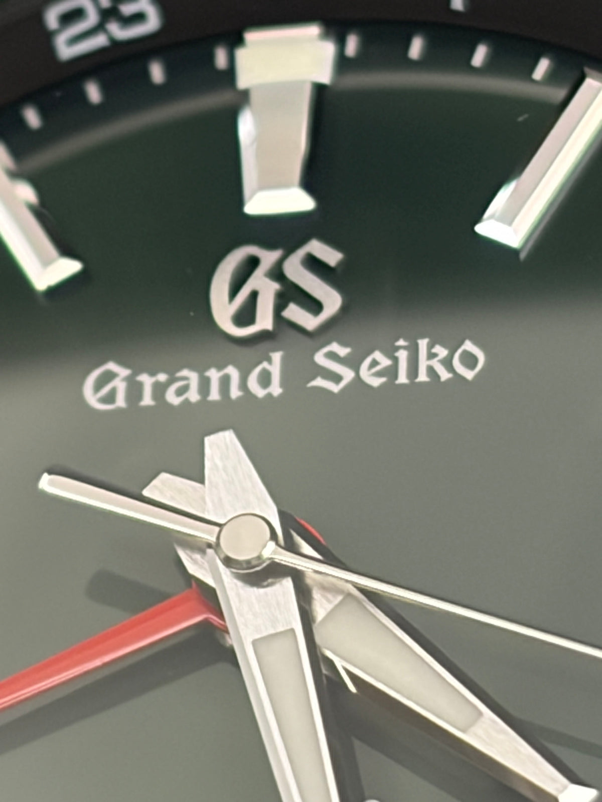 Close-up of a Grand Seiko watch dial showing the brand name, silver hour markers and hands, and a red second hand on a dark green background.