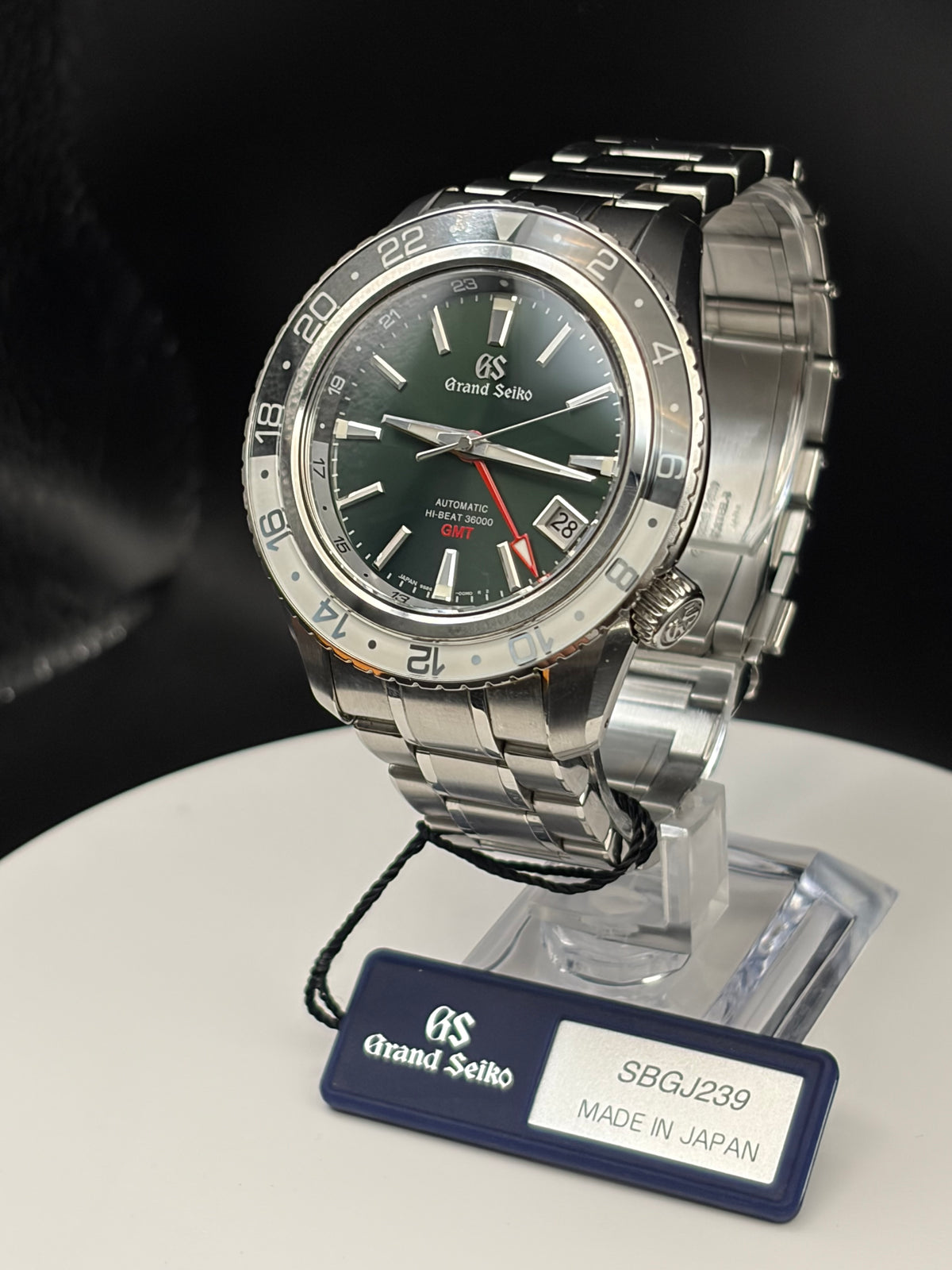 A Grand Seiko stainless steel wristwatch with a dark green dial and a silver bezel, displayed on a clear stand with a branded tag in front.