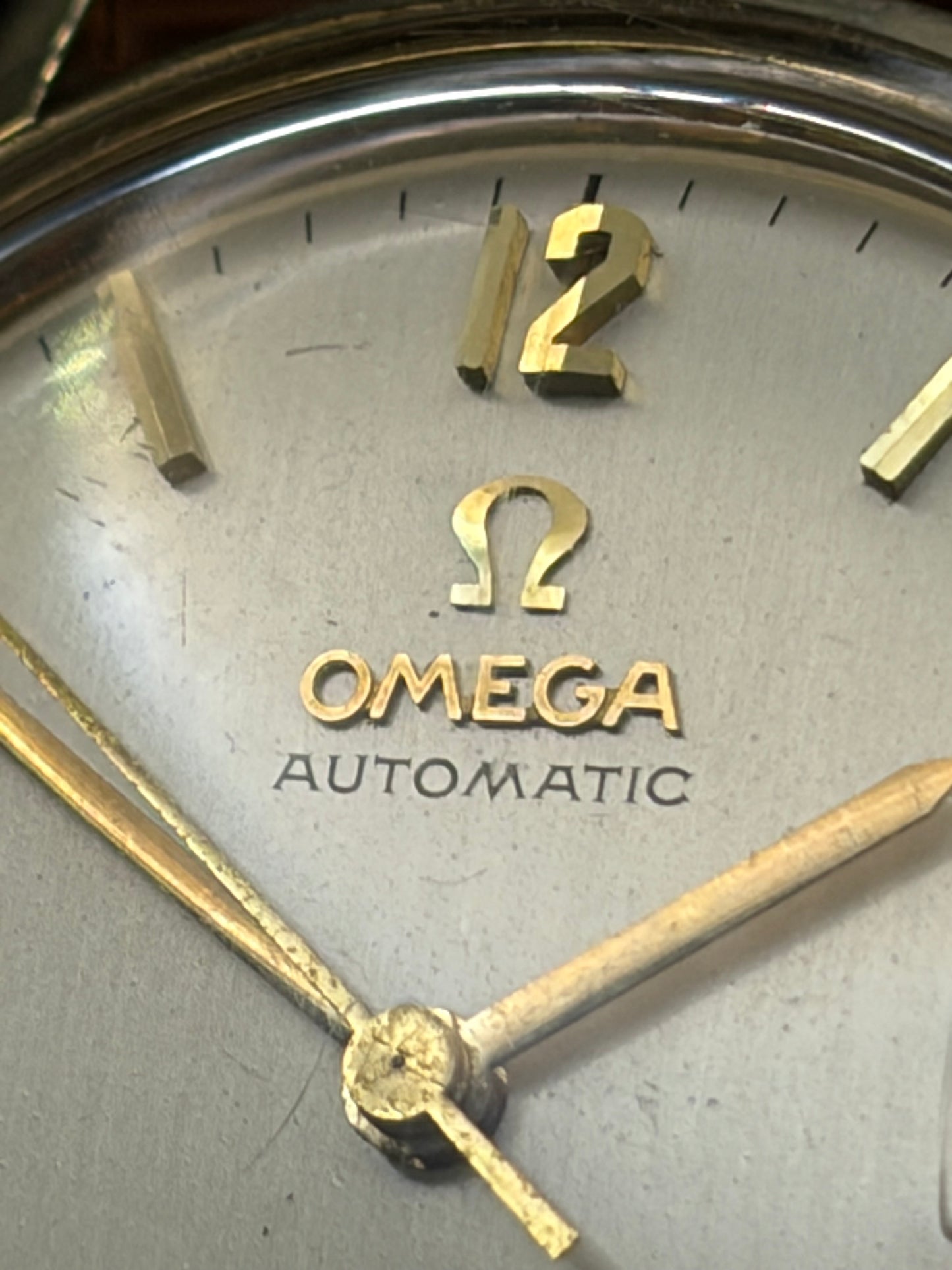 Close-up of an Omega Automatic watch dial with gold-tone hands, gold numerals, and a silver-toned textured case.