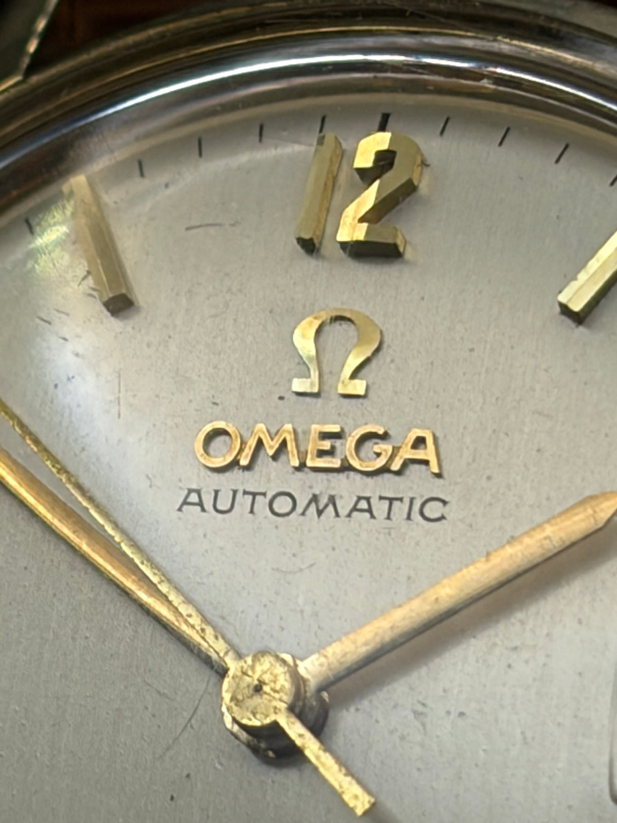 Close-up of an Omega Automatic watch dial with gold-tone hands, gold numerals, and a silver-toned textured case.
