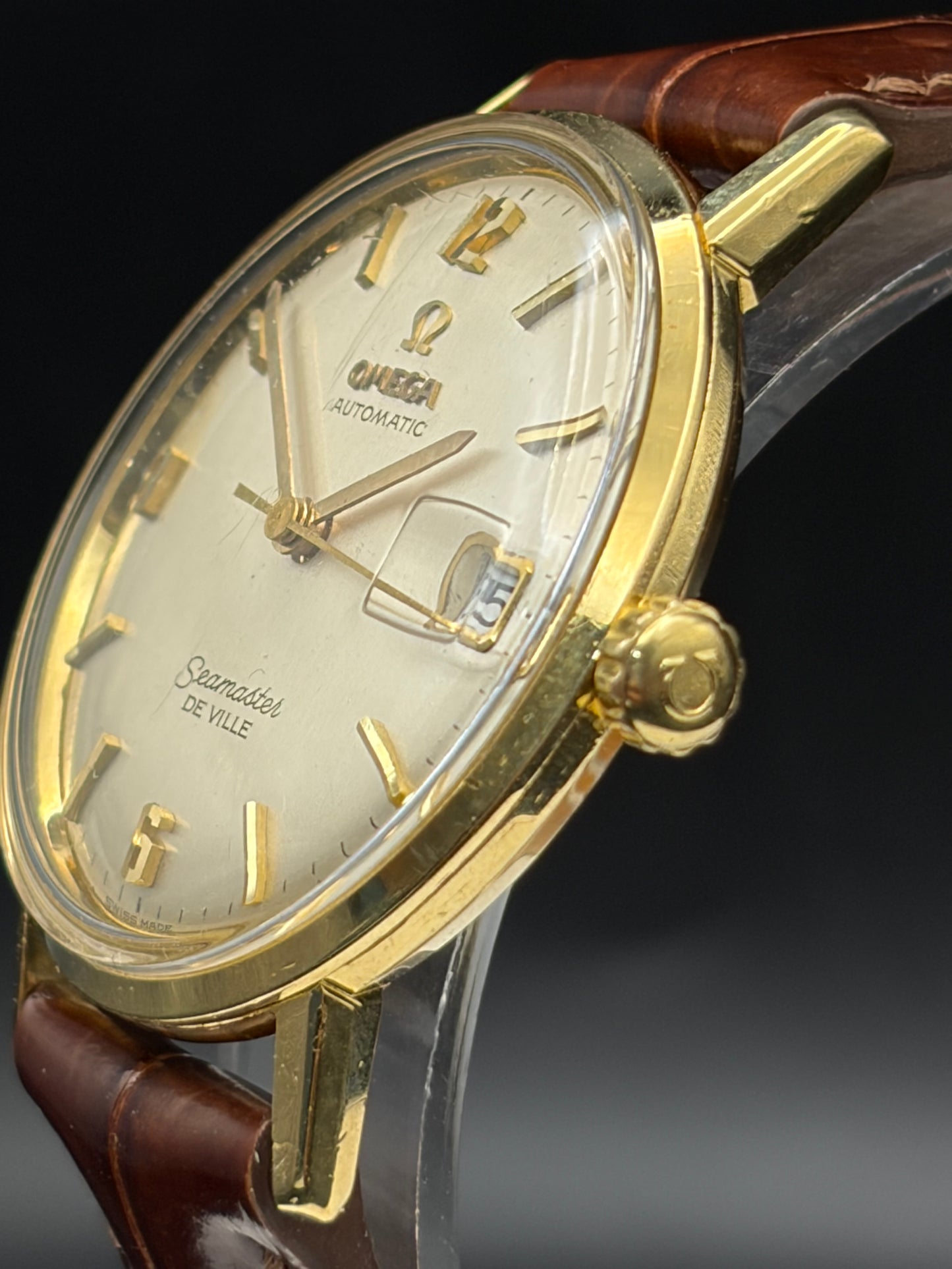 Omega automatic wristwatch with a gold case, cream dial, gold hour markers and hands, and a brown leather strap, shown in a close-up studio shot against a dark background.