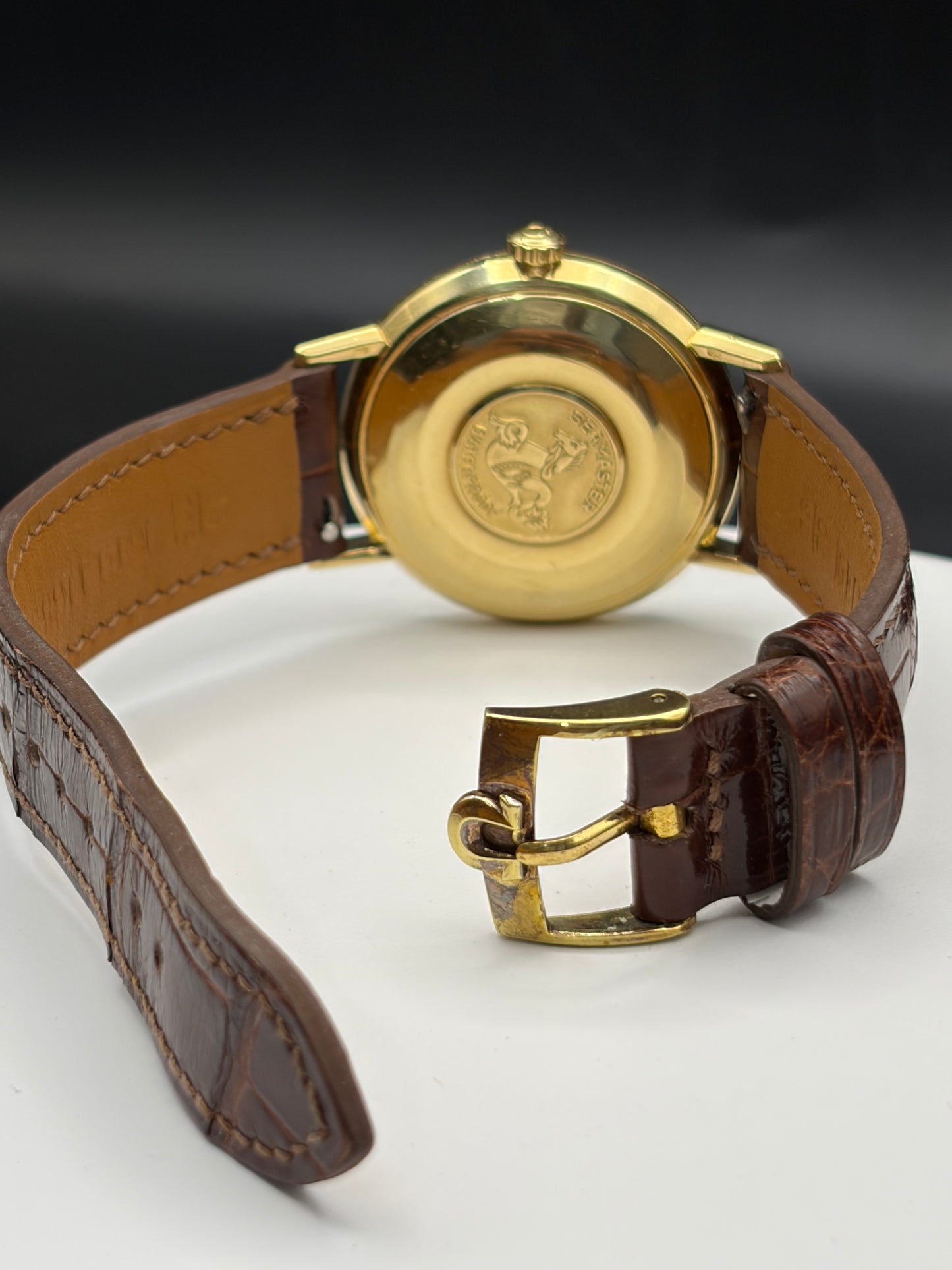 Close-up of the back of a wristwatch with a round gold-tone case, a brown leather strap, and a gold buckle on a white surface against a dark background.