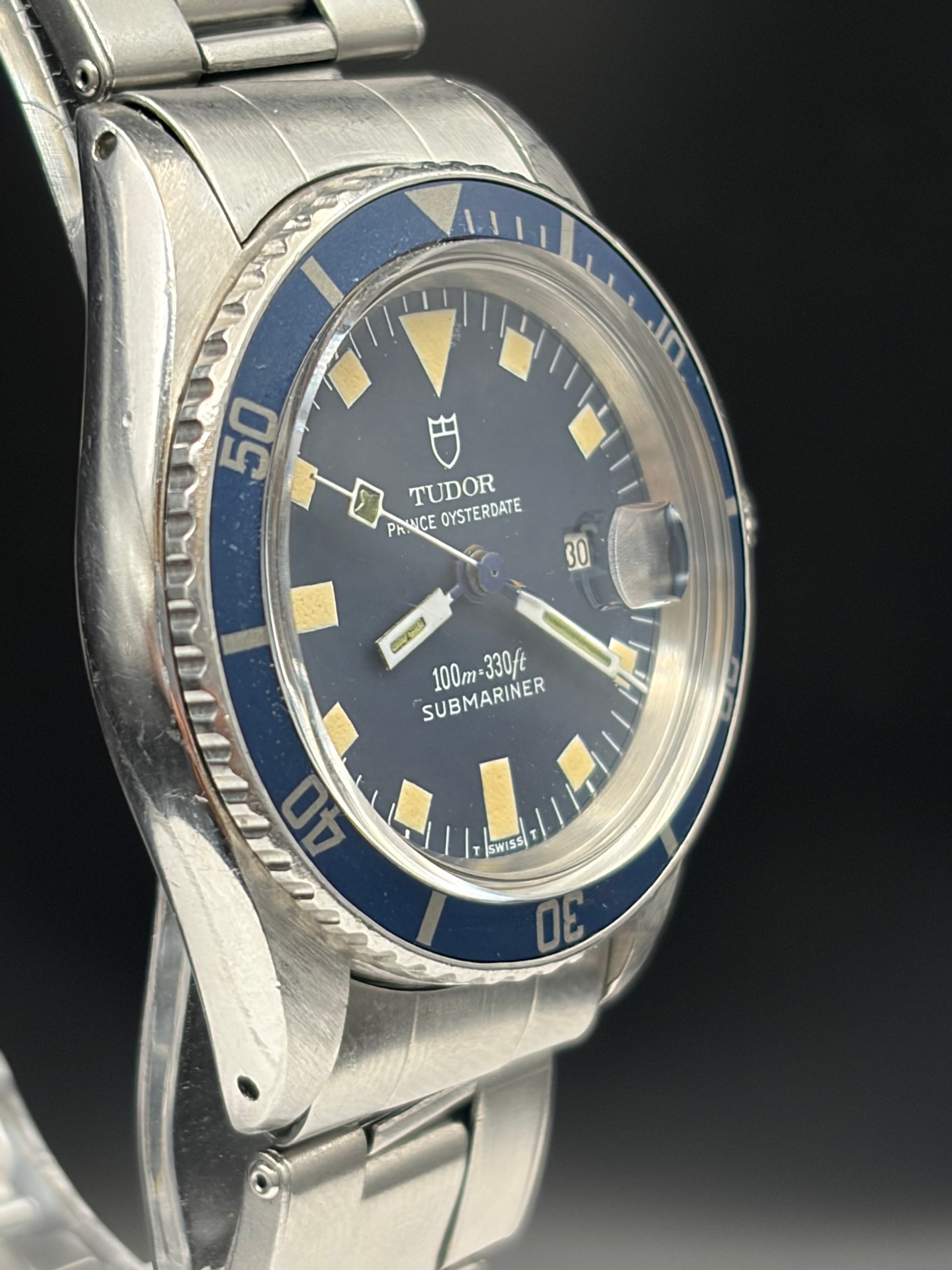 A close-up shot of a vintage Tudor Seiko Oyster Submariner watch with a stainless steel bracelet, featuring a blue dial, blue bezel, and gold-tone markers.