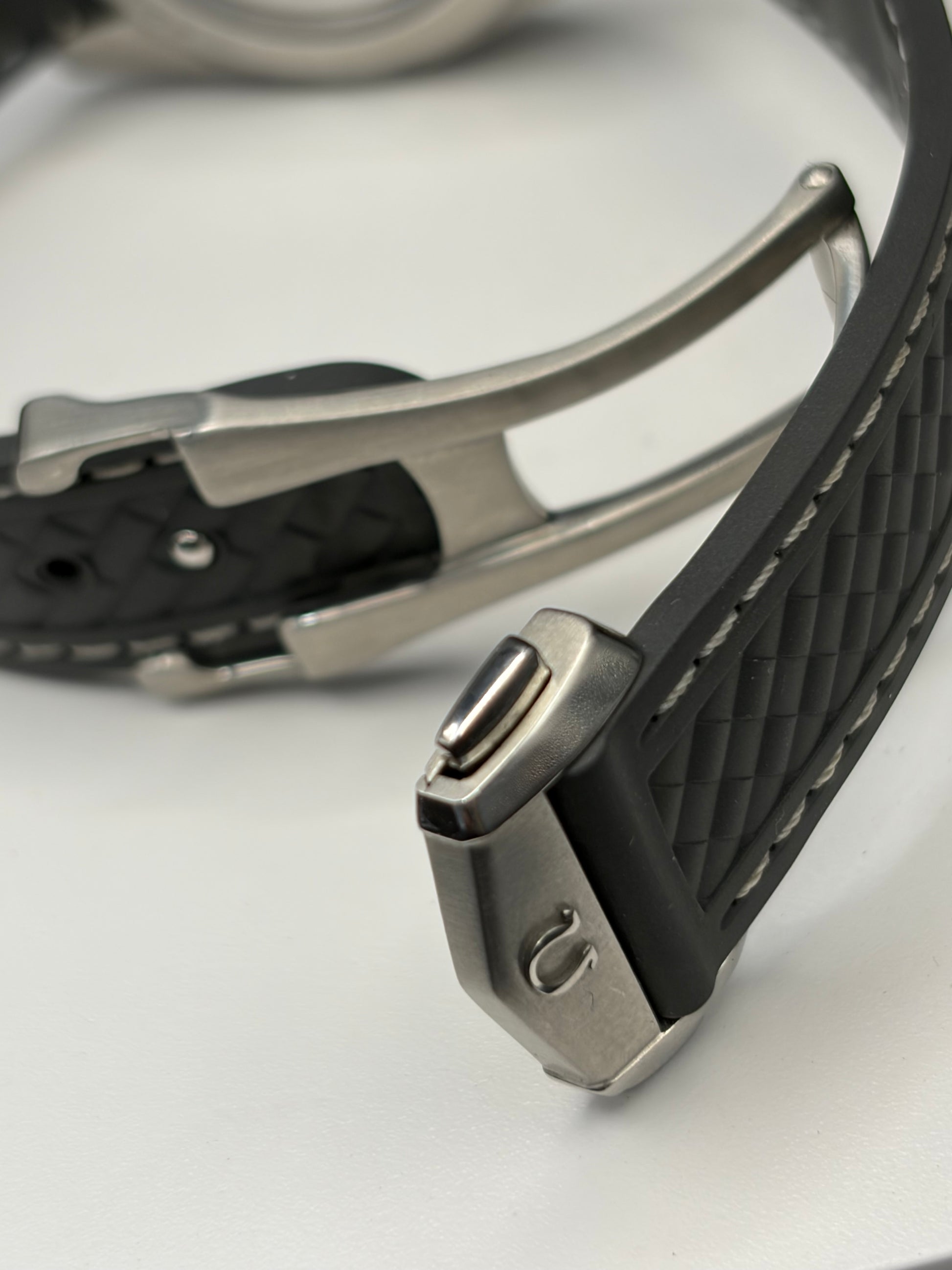 Close-up of a watch strap with a black textured silicone band and a silver-toned metal buckle on a white surface.