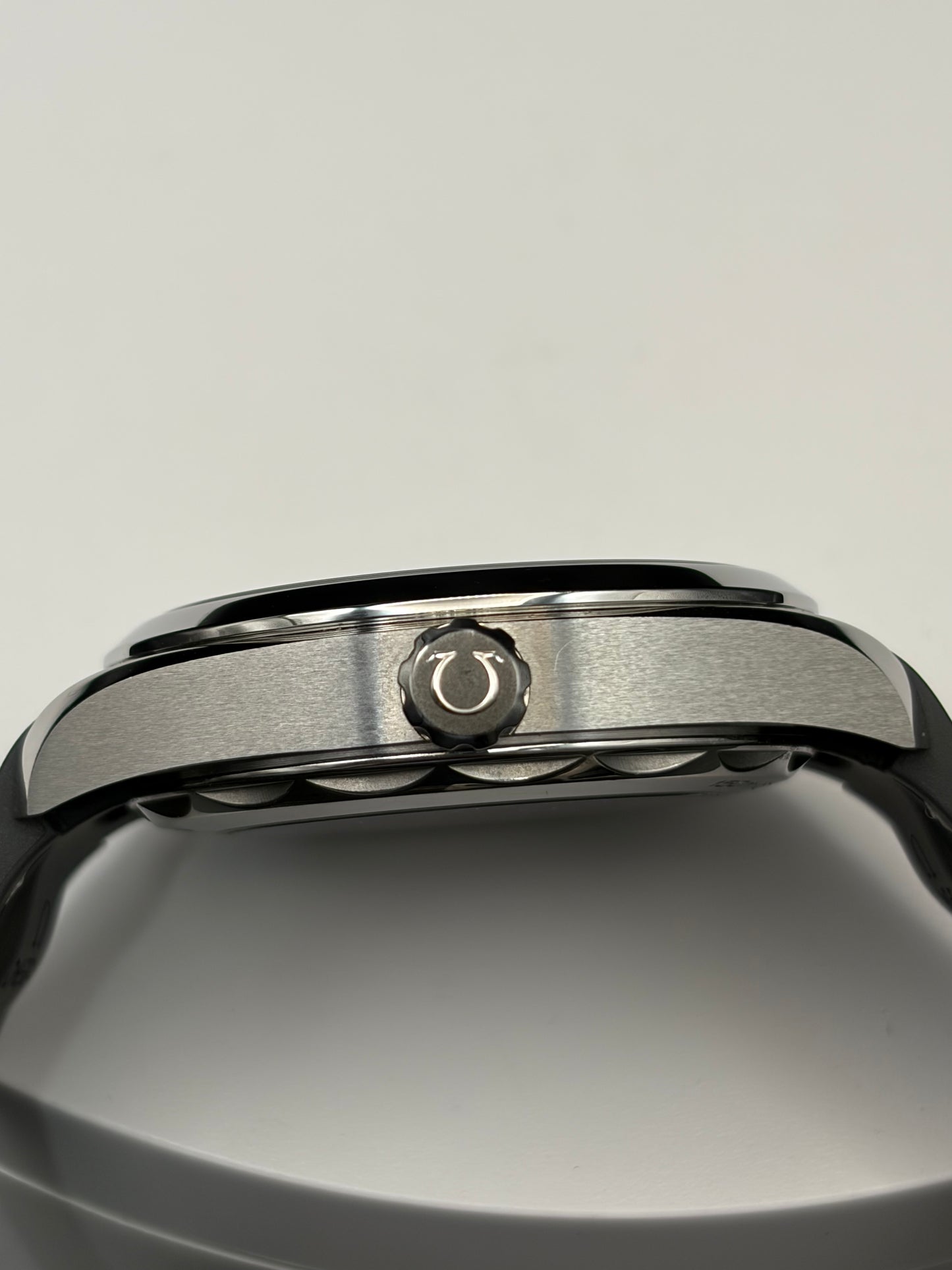 Close-up of a stainless steel watch case showing the crown and textured bezel details, resting on a neutral gray surface.