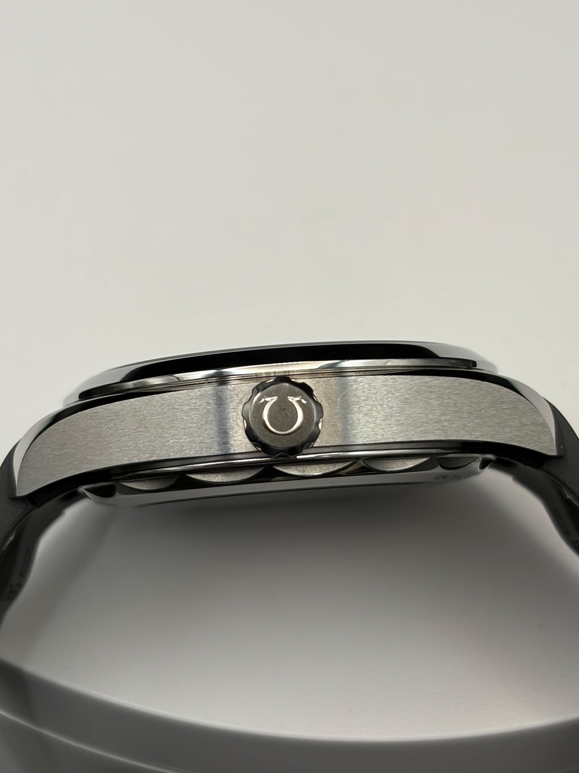 Close-up of a stainless steel watch case showing the crown and textured bezel details, resting on a neutral gray surface.