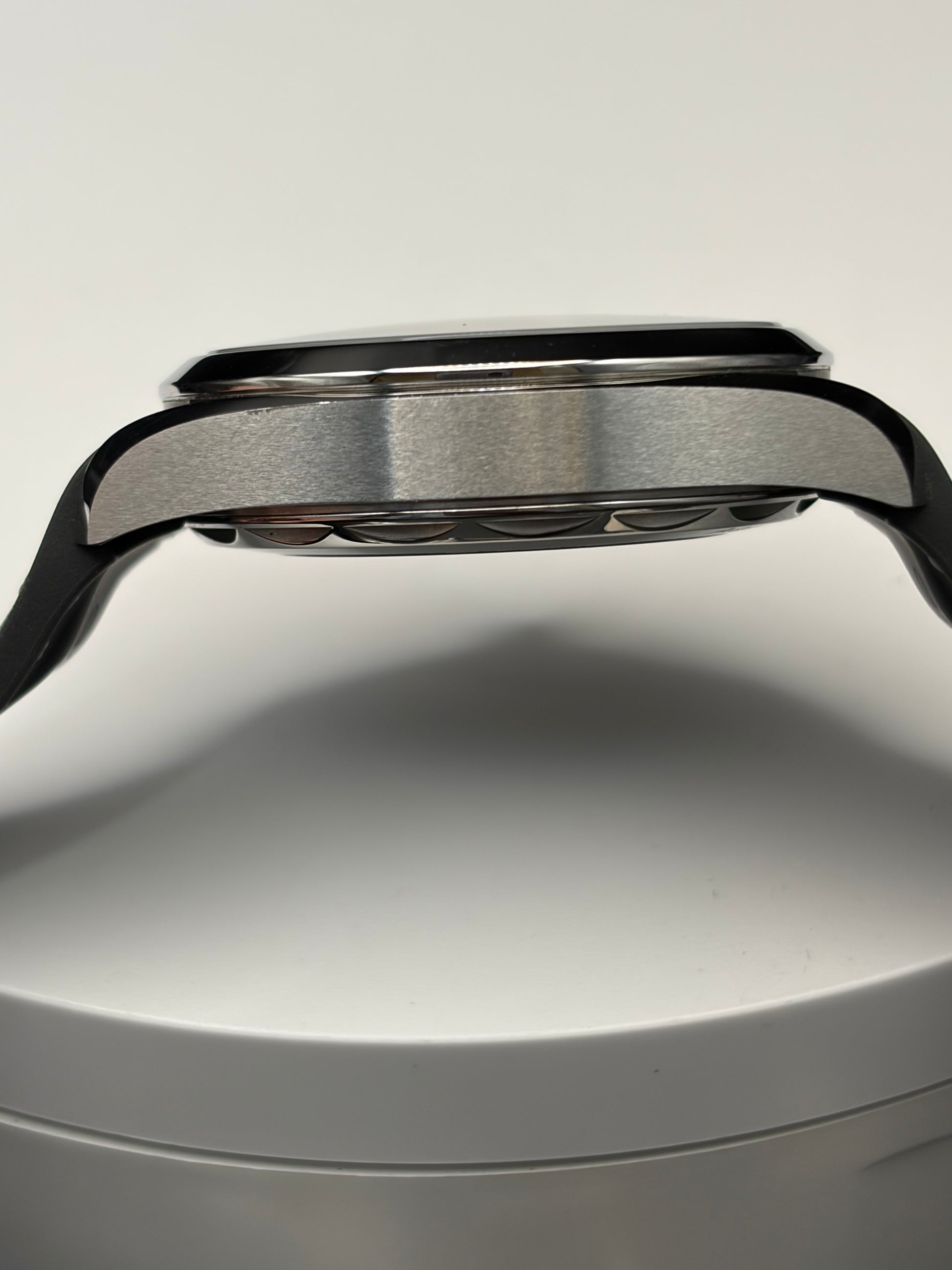 Close-up side view of a smartwatch with a brushed silver metal case and black strap, resting on a white cylindrical surface against a light gray background.