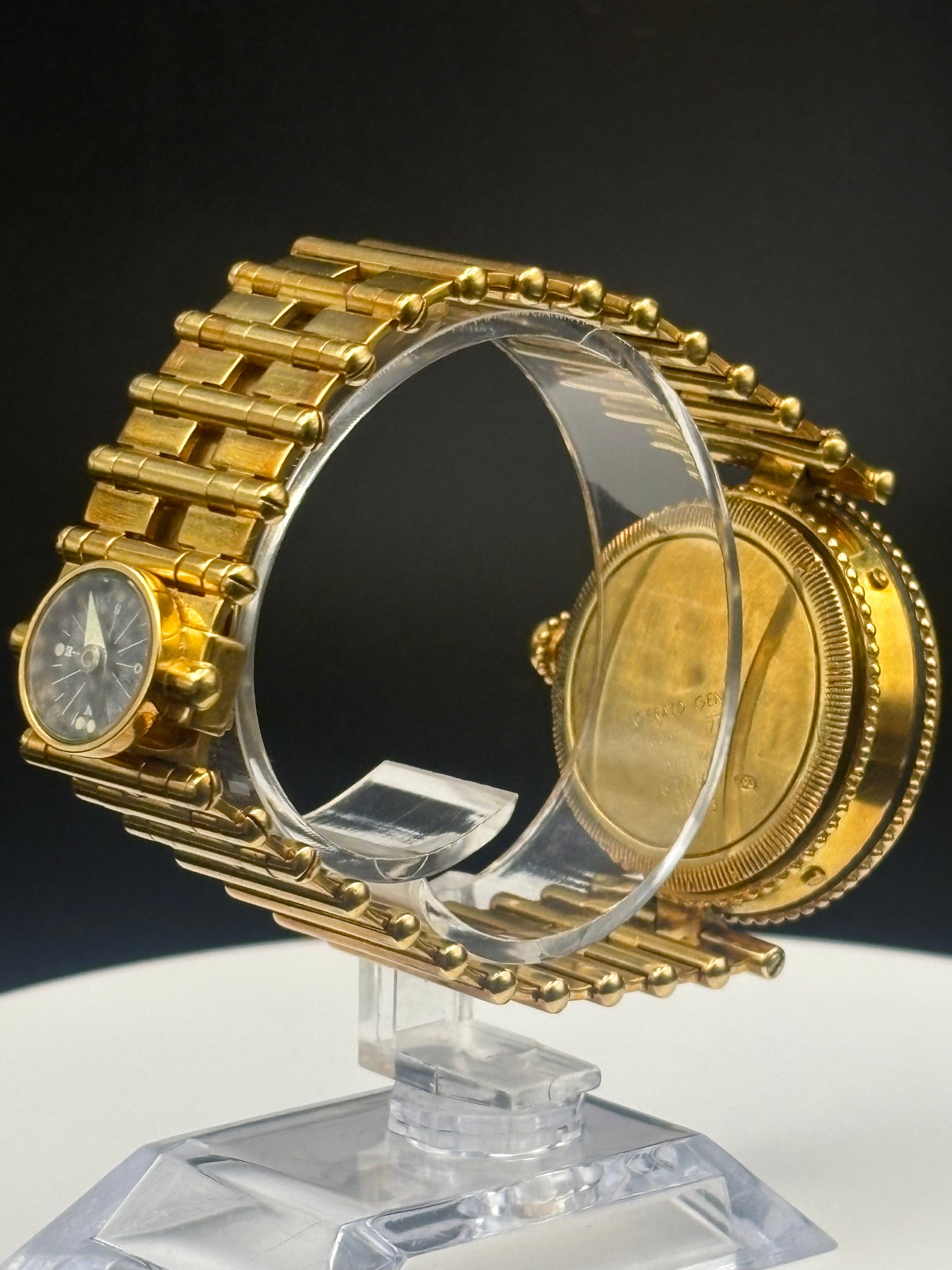 A gold-tone watch with a matching link bracelet, displayed on a clear acrylic stand against a dark background. The watch face is visible on the left, and the back casing is visible on the right.
