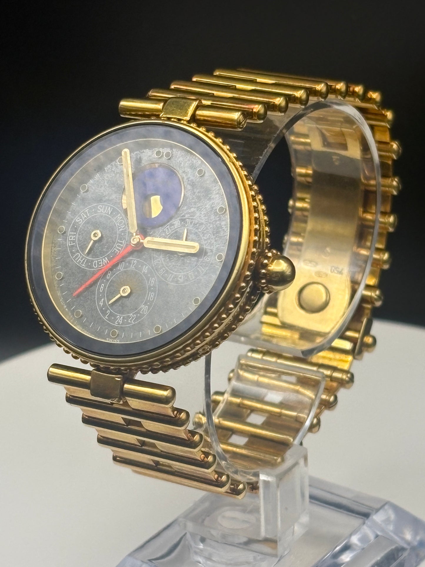 A gold-tone wristwatch with a textured gray dial, gold bezel, and gold multi-link bracelet, displayed on a clear acrylic stand against a dark background.
