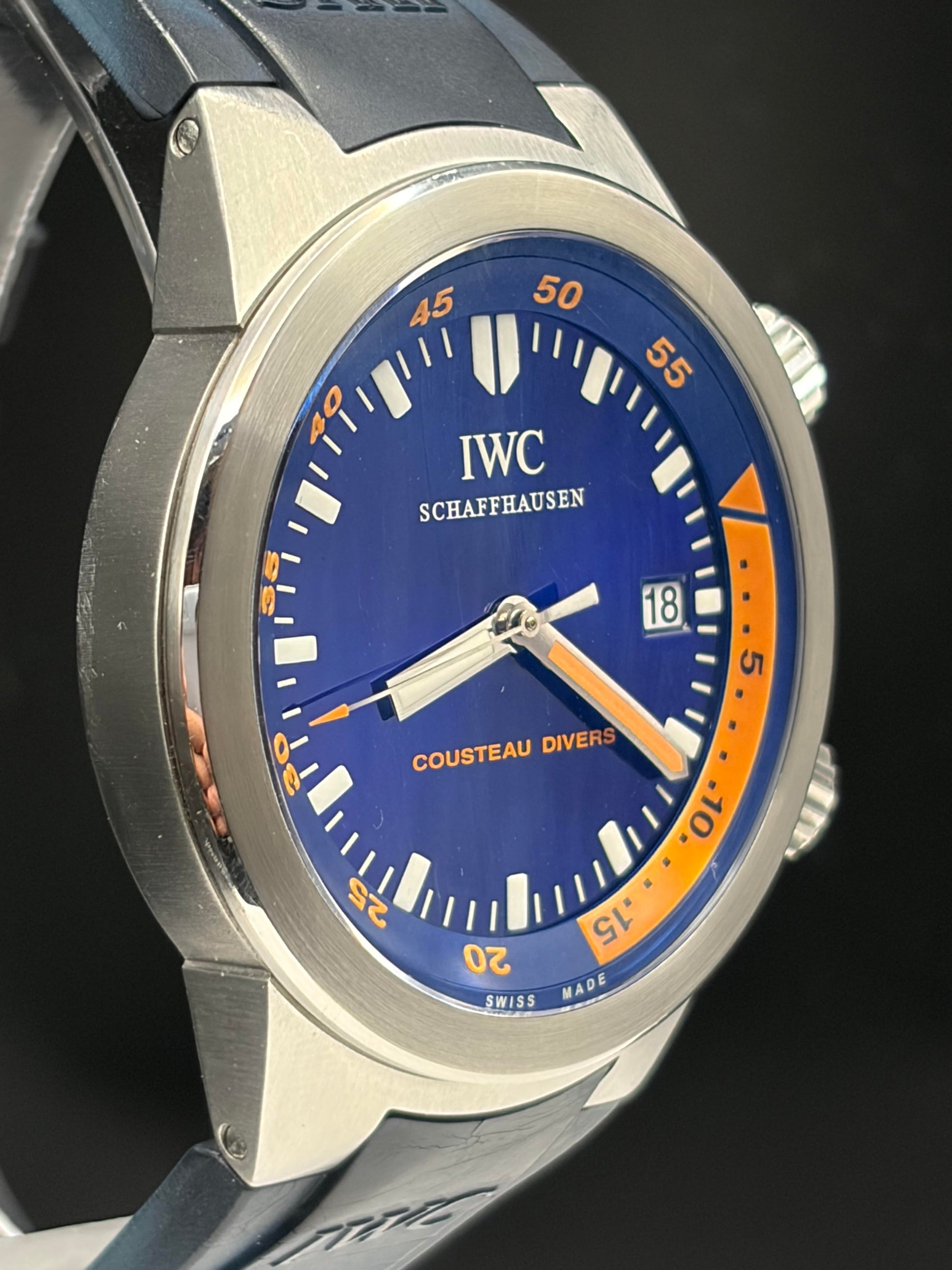 Close-up of an IWC Schaffhausen Cousteau Diver 300 wristwatch with a blue dial, orange bezel insert, silver stainless steel case, and dark gray strap against a dark background.