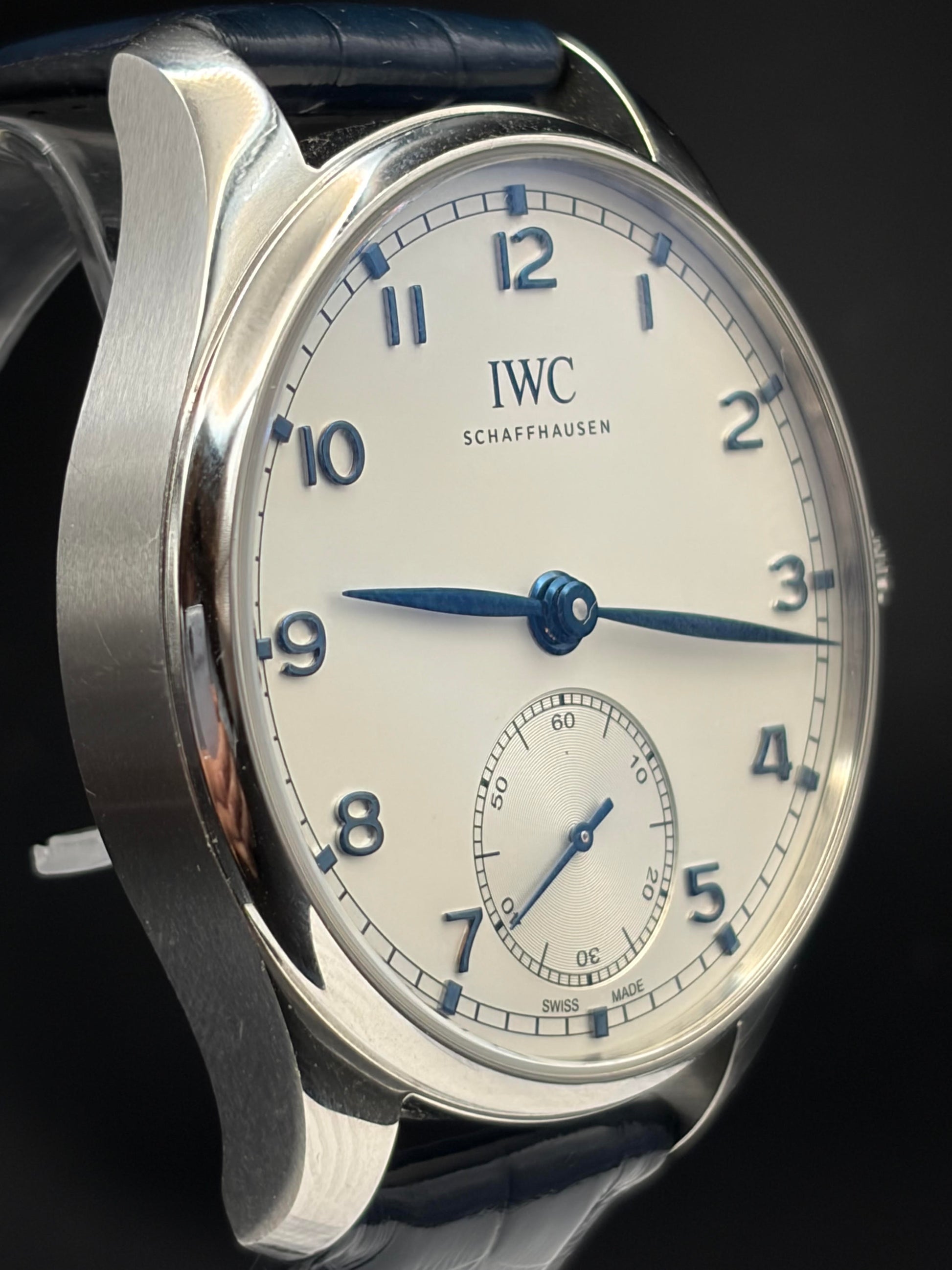 Close-up of an IWC Schaffhausen wristwatch with a silver-toned metal case, white dial, blue Arabic numerals and hands, and a dark blue leather strap against a dark background.