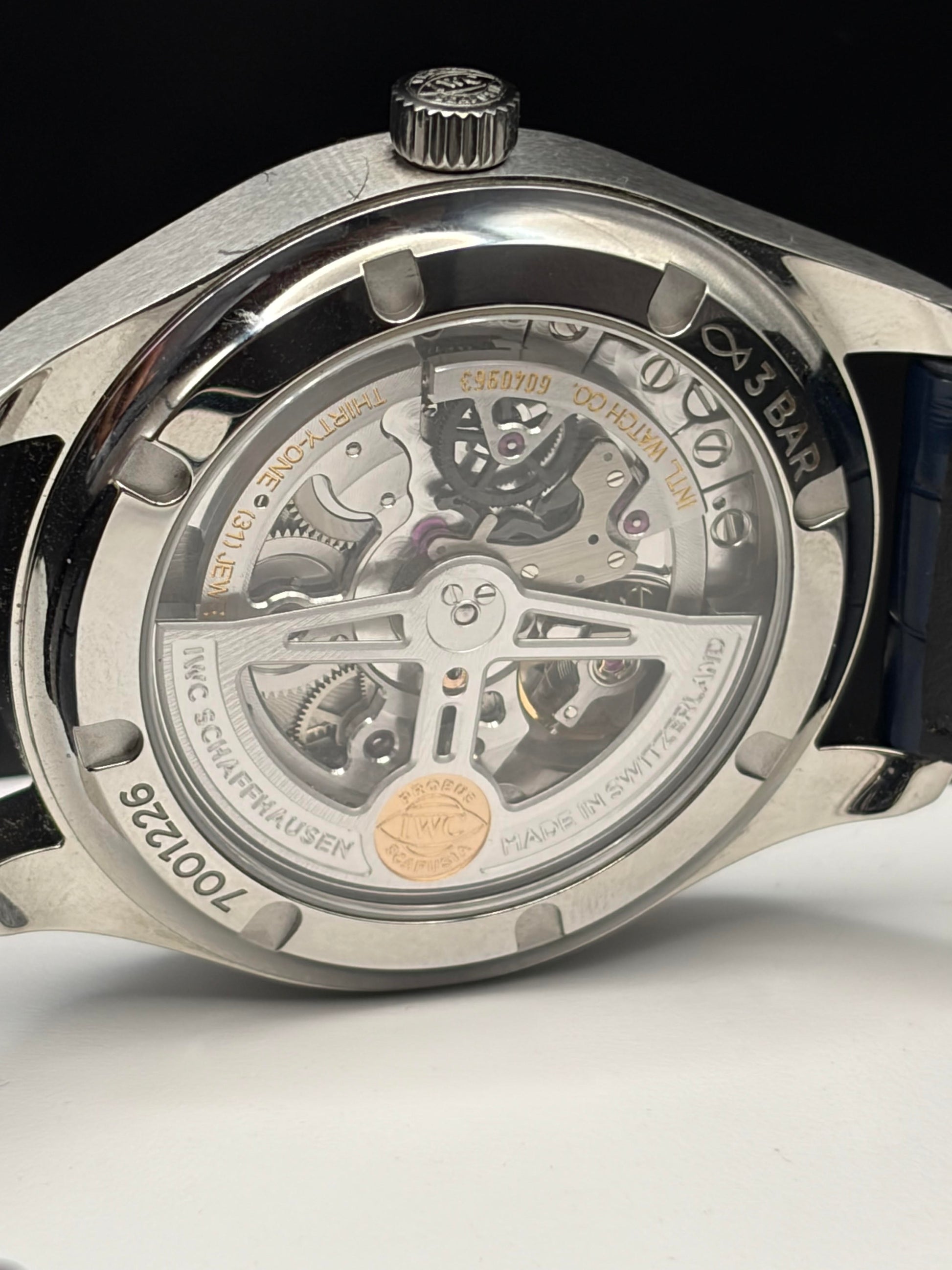 Close-up of a silver stainless steel watch back with a transparent case, showing the intricate mechanical movement inside. The watch has a dark blue strap visible on the left and a black background.