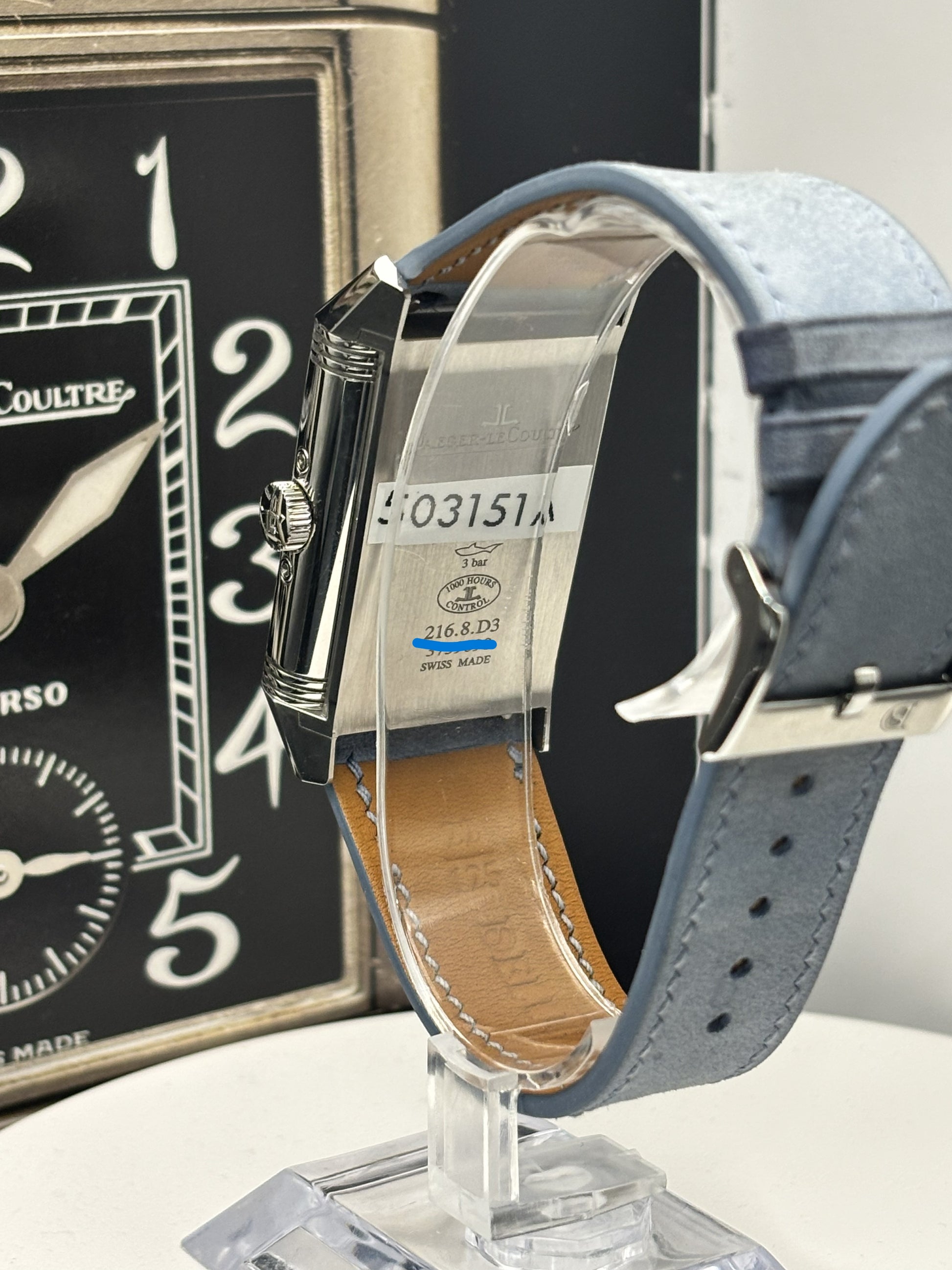 Watch with blue strap on a stand, with a clock in the background