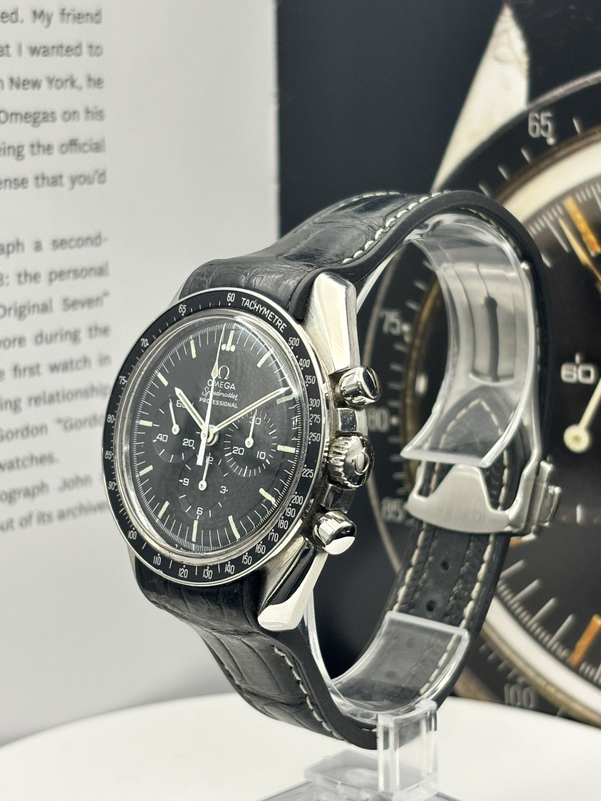 Black watch with leather strap on a stand, with another watch in the background.