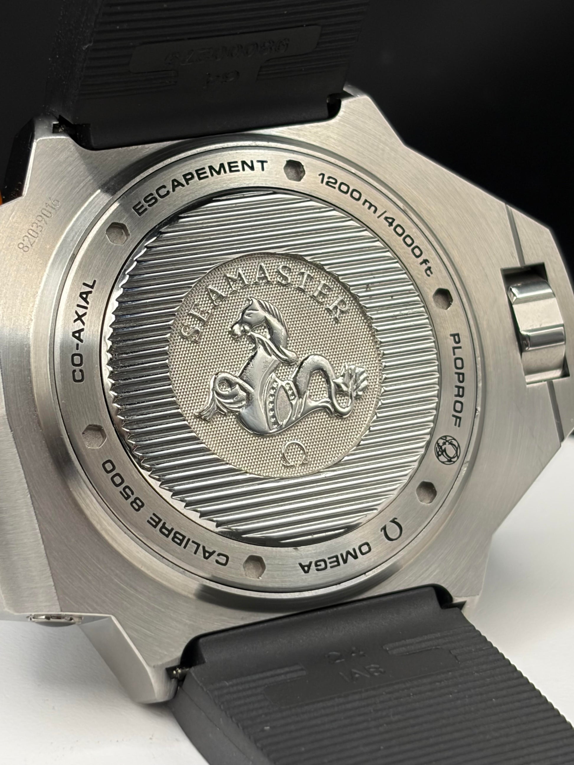 Close-up of the back of an Omega Seamaster Co-Axial watch with a brushed stainless steel case and black rubber strap, showing engraved details including the Sea Monster emblem and depth markings.
