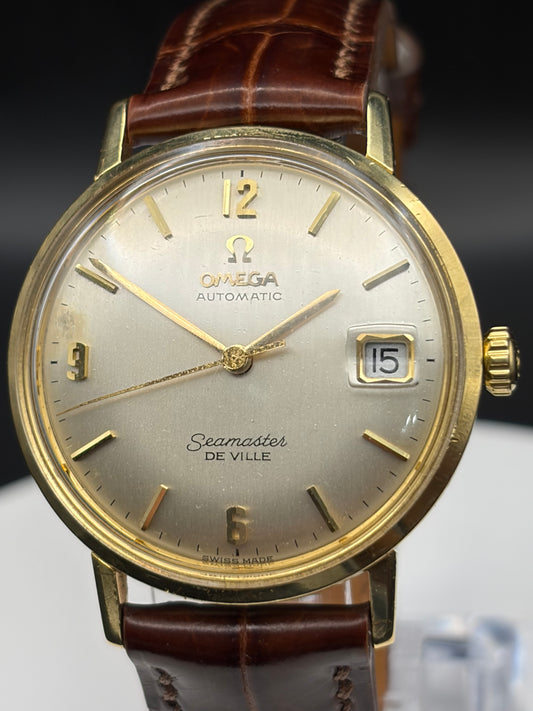 Omega Seamaster De Ville automatic wristwatch with a gold-tone case, champagne dial, and brown leather strap.