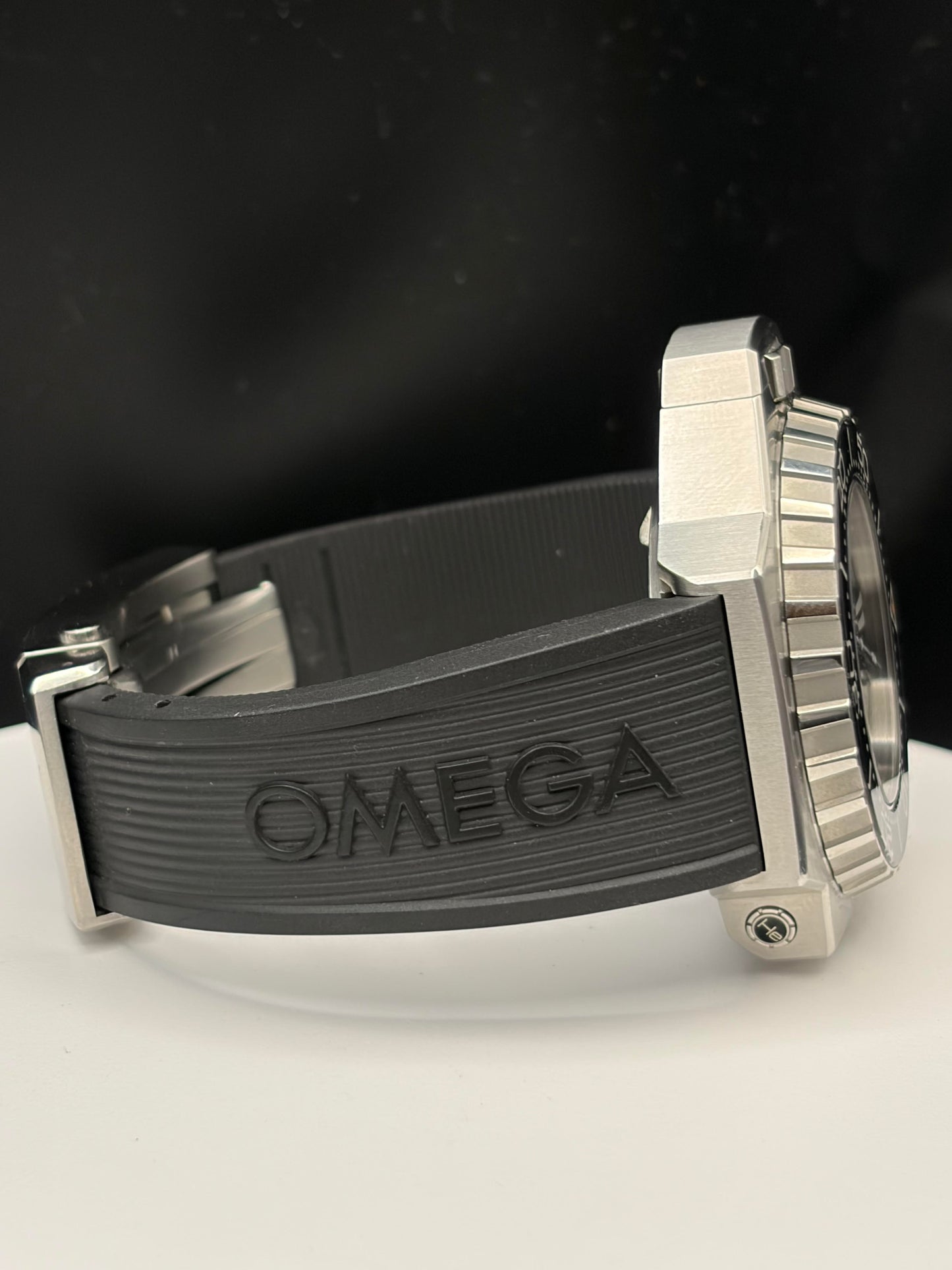 Omega watch with a silver-toned metal case and a textured black rubber strap, shown from a side angle on a white surface with a dark background.
