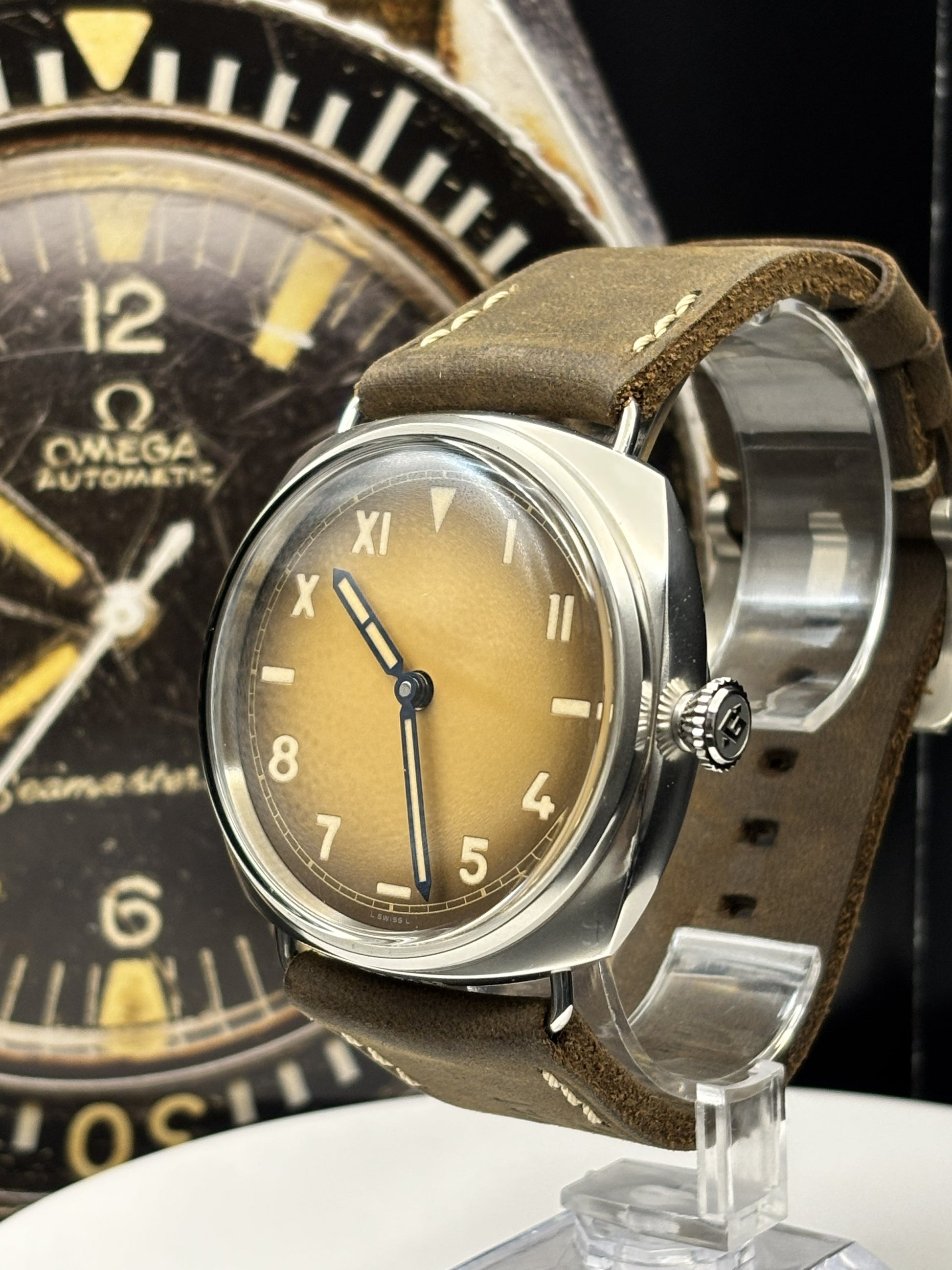 Vintage watch with brown leather strap on a clear stand, Omega watch in the background