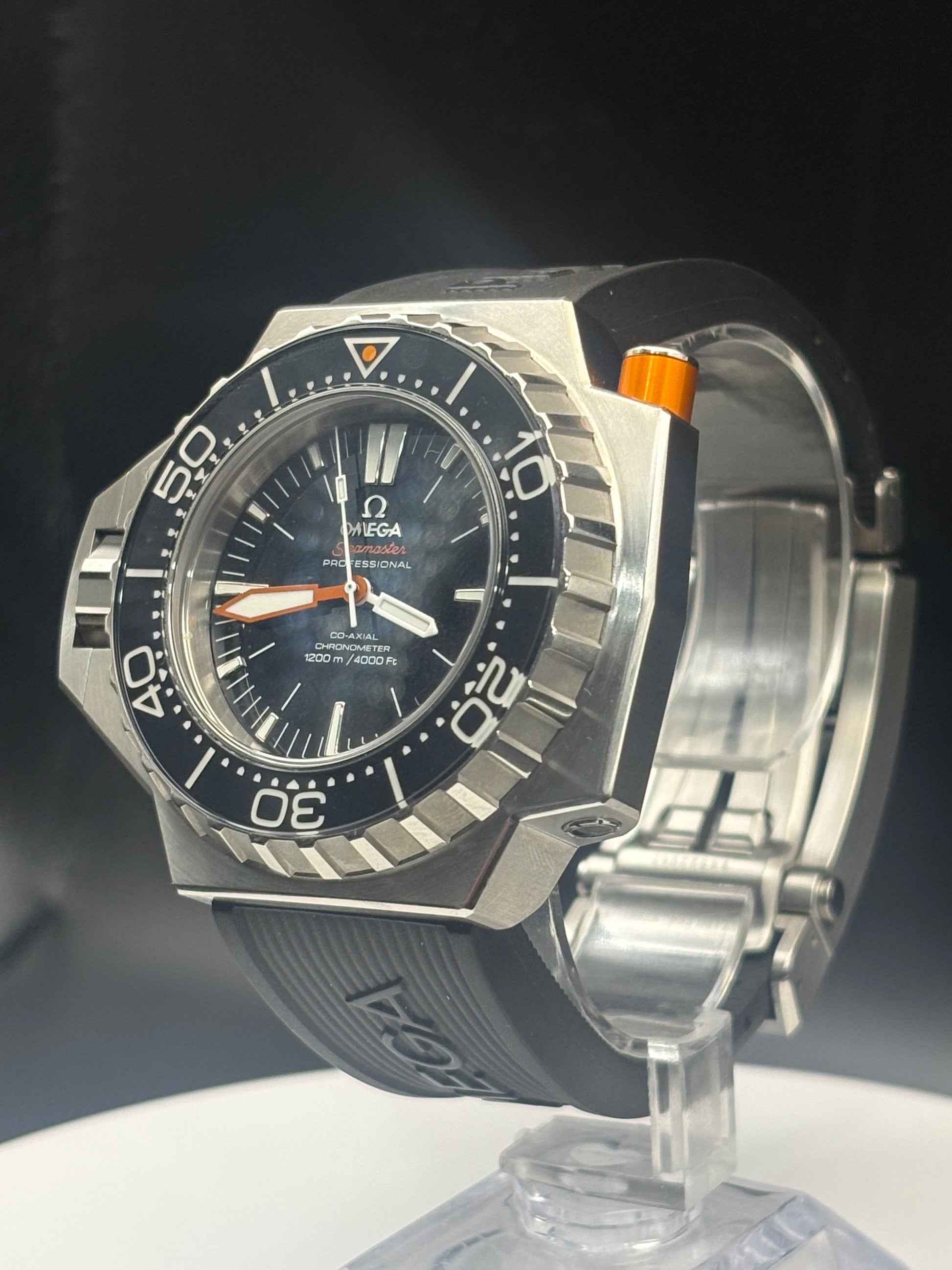 Omega x Swatch Submariner Date watch with a silver-toned octagonal case, black dial, black bezel, and black rubber strap, displayed on a clear stand against a dark background.