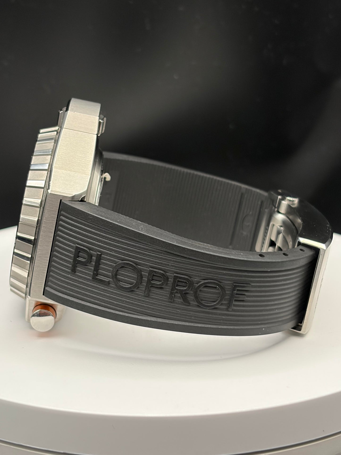 Close-up of a wristwatch with a dark gray ribbed rubber strap and a silver-toned metal case, photographed on a white circular display against a dark background. The strap is embossed with the brand name PLOPROF.