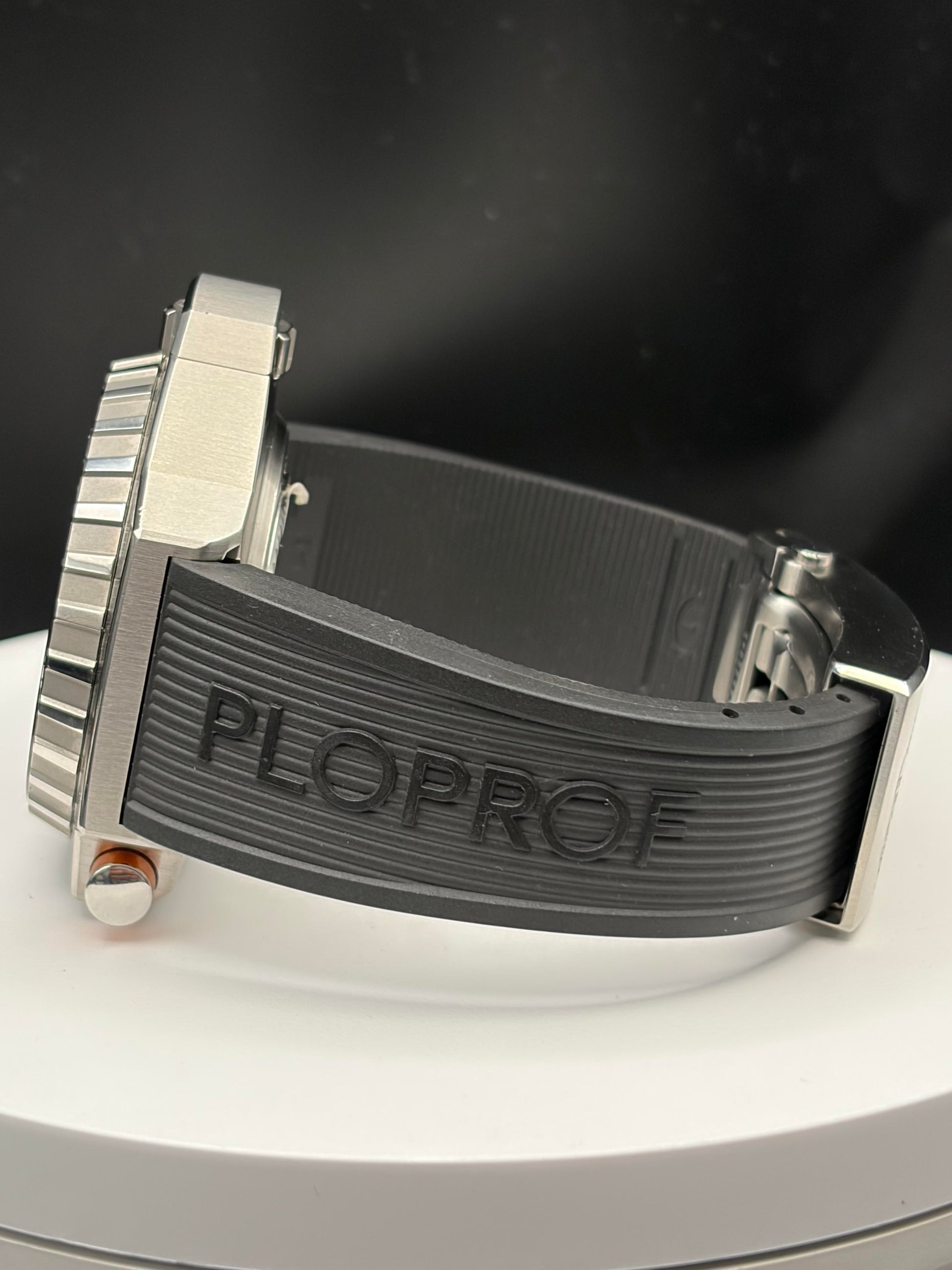 Close-up of a wristwatch with a dark gray ribbed rubber strap and a silver-toned metal case, photographed on a white circular display against a dark background. The strap is embossed with the brand name PLOPROF.